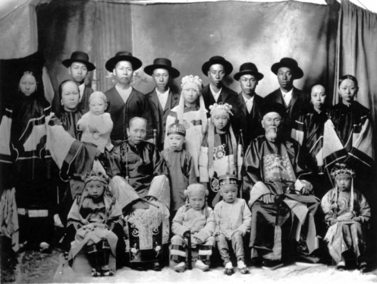 Studio portrait of a Chinese family in Idaho City, Idaho. One man wears a Manchu robe and hat. The women wear robes with wide sleeves and slippers.