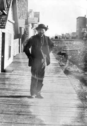 A Chinese man poses on a wooden sidewalk near a business district possibly in Colorado. A nearby sign on a building reads: "[?]ng Lee [La]undry".