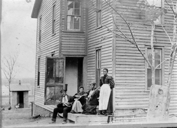 A Black man and women pose with mandolins and a guitar on the porch of a house in Denver, Colorado. A chicken and outhouse are near the two story wood frame house.