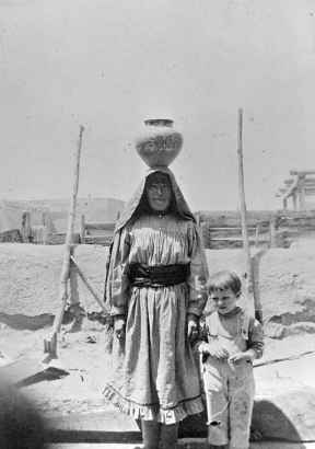 Native American (Cochiti) woman and a white boy stand in front of a pueblo wall and wire fence, Cochiti Pueblo, New Mexico. The woman carries a handcrafted pot on her head. She wears a print dress with a cloth belt and ruffled hem, and a shawl over her head. The boy wears a shirt and overalls.