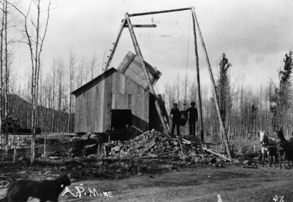 Two prospectors pose with a barrel hoist under a log frame at the A.P. Mine, possibly in Teller or Douglas County, Colorado; shows wood plank structure with open door on roof and opening for an ore car on side, rock pilings, team of horses, and a dog.