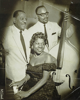 Three entertainers pose for a group photograph. Two unidentified males wear suites and ties and one plays the cello. Denver native and jazz pianist, Charlotte Mosley Cowens, sits at the piano wearing an off-the-shoulder dress and diamond jewlery.