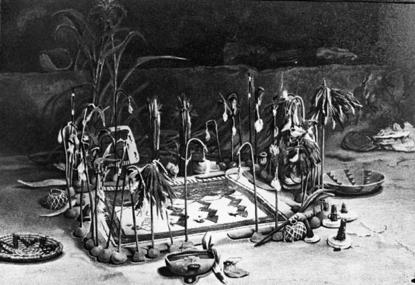 Native American (Hopi) antelope altar shows sandpainting; clay pedestals and crooks (ceremonial bows) around painting, water gourds, corn stalks, baskets, medicine bowl, rattles; Walpi pueblo, Arizona.