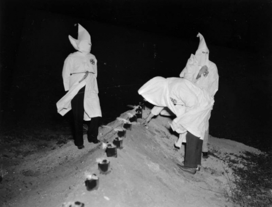 Ku Klux Klan memeber light candles in metal cans buried in a dirt mound probably in Denver, Colorado. They wear hoods and robes.