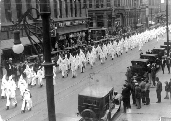 Members of the Ku Klux Klan march in a parade on 17th (Seventeenth) Street in Denver, Colorado. They wear hoods and robes as spectators look on. Parked automobiles line the street. A sign on a building reads: "Western Clothing Co."