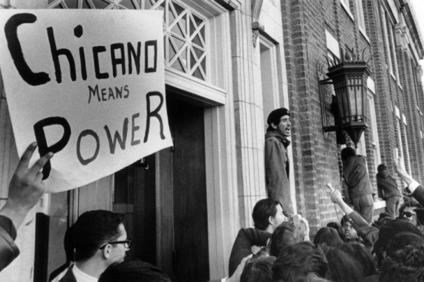 A Mexican-American man speaks into a microphone in Denver, Colorado, during a protest. Picket sign reads: "Chicano Means Power." An ornate lamp is on a brick building with entablature.