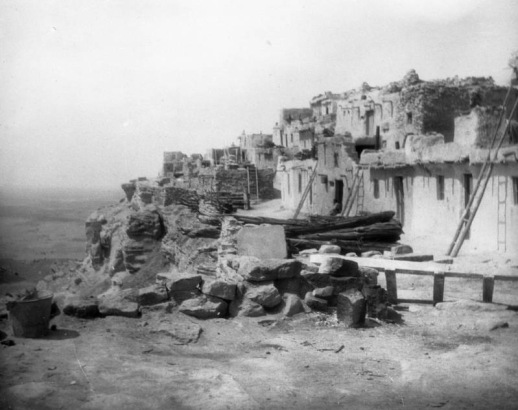 Native American (Hopi) Walpi Pueblo, Arizona with multi-story adobe and masonry structures on mesa with windows, doors, and ladders. Shows wood pile, metal bucket, ladder against a stone wall in foreground, valley in background.