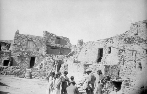 Native American (Hopi) men and children stand in a courtyard at Oraibi Pueblo, Third Mesa, Arizona. One man in the group holds a basket. The adobe and stone cluster homes have adobe stairs, wooden vigas, and wood pole cross beams supported by the walls. A tripod stands in the courtyard.
