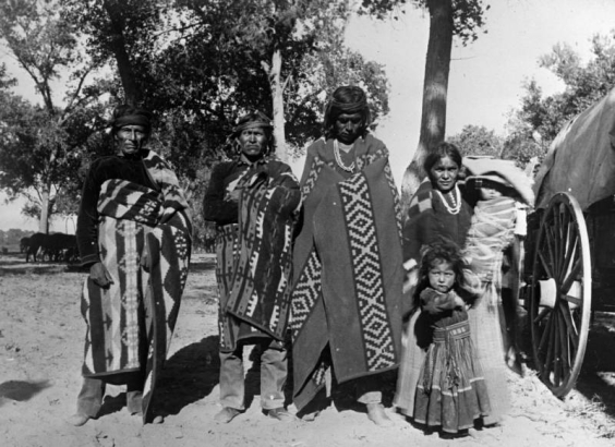 A group of Native American (Navajo) men, a woman, child, and baby in a cradleboard pose outdoors near an unhitched wagon at Shiprock, New Mexico. The men are wrapped in woven blankets and wear headbands. The woman wears a dress and shawl. The girl wears a dress with a wide belt. The woman holds a baby in a cradleboard.