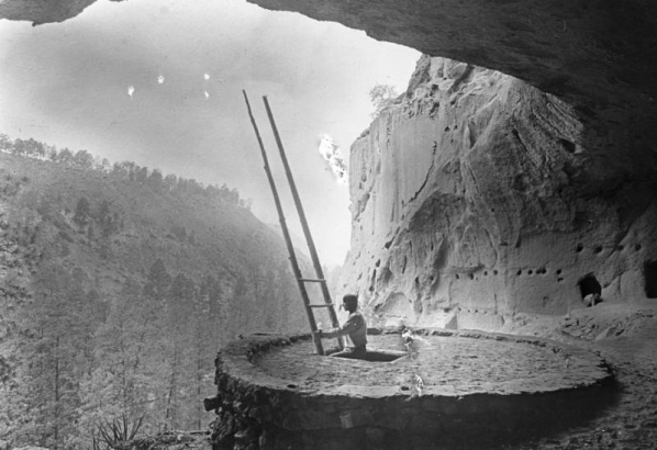 A Native American (tribe unknown) man stands on a wooden ladder which leads out of a kiva at Ceremonial Cave, near Frijoles Canyon, Bandelier National Monument, New Mexico. The round, covered kiva stands in a cliffside cave.
