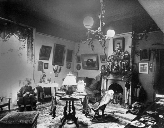 Interior view of the Drawing Room at the Palace of Governors, Santa Fe, New Mexico. Chairs, tables, a footstool, and a lamp sit in the center of the room. A boy sits in one of the chairs that line the wall. A couch sits next to the fireplace. Paintings are hung on the walls. Greenery hangs from the chandelier. Statuettes stand on the mantle.