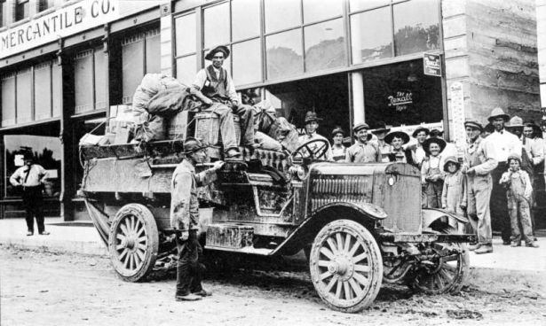 A group of Native American (Navajo) men and boys, in hats, shirts, overalls or pants,  pose on a sidewalk in Farmington, New Mexico, by the Farmington to Shiprock auto truck. The convertible truck has packages and bundles tied to the back and one man seated on a square basket; another man stands by the running board. Shows store fronts with signs that read: "Mercantile Co." and "The Rexall Store" and a large "Rexall" thermometer on wall.