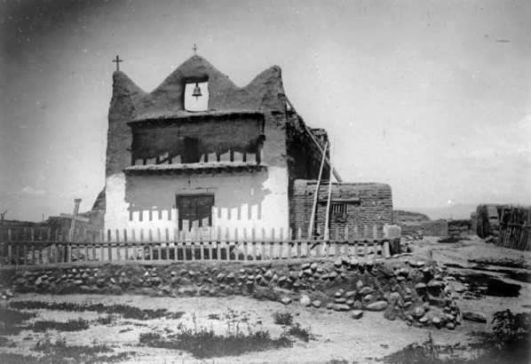 Church at Native American (Tewa) Nambe Pueblo, New Mexico; an adobe mission with double wood doors, three point facade and center belfry, balcony, attached structure and a picket fenced courtyard.