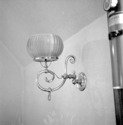 Interior view of the Hamill House, in Georgetown, Colorado. The house was built by Joseph Watson, and was acquired by William A. Hamill, his brother-in-law, in 1874. Between 1874 and 1881 Hamill made various additions to the house and grounds. The image shows a metallic light fixture attached to a wall, with a curvilinear stem with a gas regulator which supports a textured glass globe. The wall and ceiling have subtly textured wallpaper.