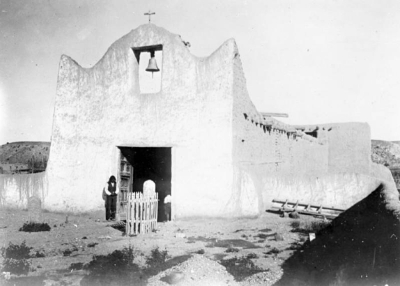 Church at Native American (Tewa) Santa Clara Pueblo, New Mexico, southwest view. Shows adobe mission with a scalloped facade and center belfry; gravemarkers in courtyard. Native American (Santa Clara Pueblo) man stands by the church entrance. He wears a shirt, pants, and vest.