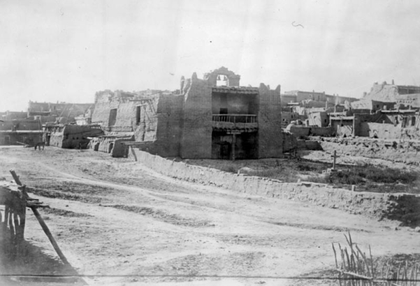 View of Mission Nuestra Senora de Guadalupe in Zuni Pueblo, New Mexico. Shows an adobe mission with a belfry and two bells, a balcony with railing over the entrance, a center support post under the balcony, vigas, and walled courtyard. Multi-story pueblo dwellings are in the distance.