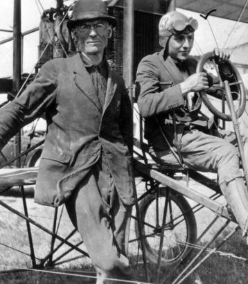 Portrait of Will "Billy" Parker, pilot, at the wheel of a 50 horse power 2 cycle Roberts engine airplane; he wears goggles and a helmet lettered: "13." P. B. Belt stands alongside.