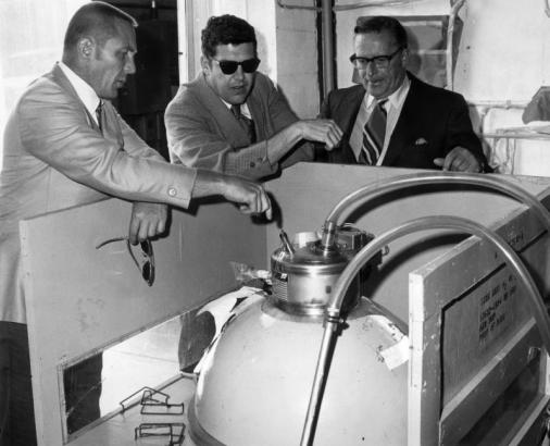 John "Jack" L. Swigert, pilot, astronaut, and public servant, points at a cryogenic oxygen tank at Beech Aircraft, Boulder Division, Boulder, Colorado; Keith E. Helart (sunglasses) and Howard E. Mers look on.