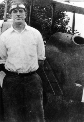 Louis Paul "Gus" Quinn, pilot and educator, poses by an airplane; his outfit includes goggles and a flight cap.