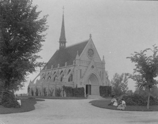 Shows the Fairmount Cemetery's Little Ivy Chapel, located in Denver, Colorado. The chapel features a compound arch doorway, wheel window, and a spire possibly with a bell tower. Two women and a child sit on the front lawn. The church opened in 1890.
