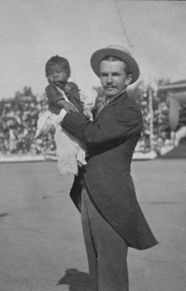 A man poses with a Native American (tribe unknown) infant in the stadium at the Festival of Mountain and Plain. The baby is wearing an oversized shirt and tattered pants. The man is wearing a suit and hat.