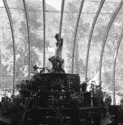 Interior view of the solarium of the Hamill House, in Georgetown,  Colorado. The house was built by Joseph Watson and acquired by mine owner William A. Hamill in 1874; shows the pyramidally tiered cast iron fernery, or plant stand, in the foreground, with potted plants on the shelves. A pewter sculpture of a nude boy blowing a horn is at the top of the stand. The decorative ribs of the solarium are behind; foliage of the trees outside shows through the glass.