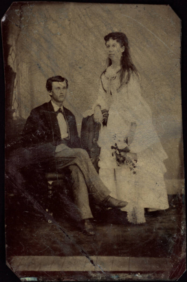 Studio portrait (sitting and standing) of a husband and wife probably on their wedding day. She holds a bouquet and wears a dress with a bustle.