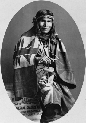 Studio portrait (sitting) of a Native American (Navajo) man. The man wears a woven blanket and holds a stone and wood hand drill. He sits on a wooden crate that reads: "[?] Factory Hartford [G]eneral Sales of [?] 253 Broadway [Ne]w York, U. S. A.".