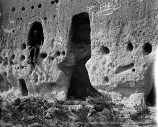 Man sits in a window opening of a Native American (Tewa) talus dwelling at the Puye Cliff Dwellings, Santa Clara Pueblo, New Mexico. The talus dwellings have holes for vigas and petroglyphs on the exterior wall.