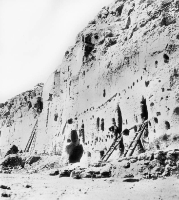 A man stands on a wooden ladder at the Long House Ruin, Native American (Anasazi) talus dwelling, Bandelier National Monument, New Mexico. Other ladders lean against the tuff dwellings which have holes for vigas.