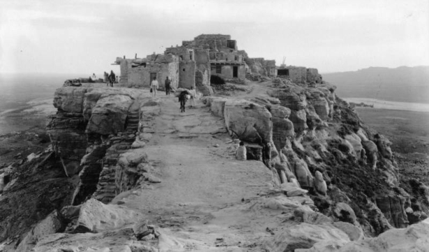View of Walpi, Arizona, a Native American (Hopi) stone and adobe pueblo settlement; shows people, a burro, and the sandstone cliffs of the mesa.