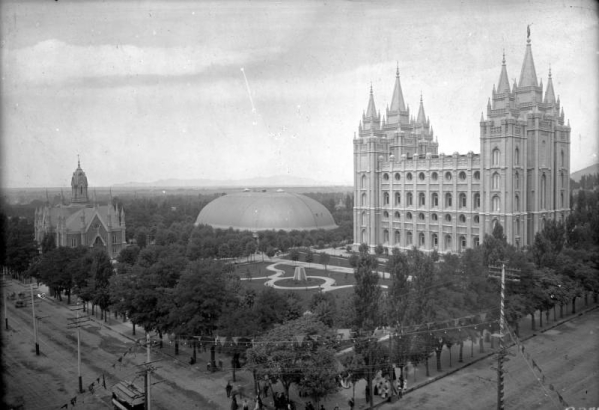 View, facing northwest at the corner of Main and South Temple Streets, of Temple Square, in Salt Lake City, Utah. The Temple of the Church of Jesus Christ of Latter-Day Saints, the Mormon Tabernacle, the Seagull Monument, and Assembly Hall (Endowment House) are in view. A gold statue of Moroni on the main spire of the temple is visible. A trolley car, tracks, and electrical wires are on and above South Temple Street.