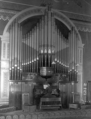 View of pipe organ behind a wood railing in St. James Methodist Chruch in Central City, Colorado.  A woman in a patterned dress poses on the bench; two wooden chairs are opposite. A sconce is attached to each side of panneling above the organ keyboard.  The floor is carpeted, and the wall around the organ is painted with decorative elements, including two columns with doric capitals.  Dedicated in 1899, the organ cost $3,000.00. to build and was water-powered until 1932.