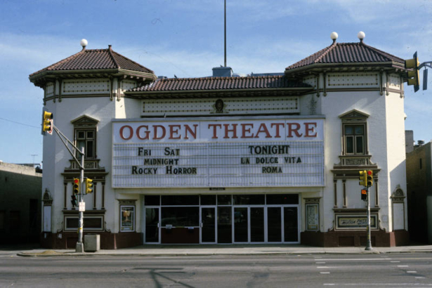View of the Ogden Theatre (1917; Harry Edbrooke) at 935 East Colfax in the North Capitol Hill neighborhood of Denver, Colorado. Shows the two-story Mediterranean Revival-style theater.  The walls of the theater are brick and the roof is covered in red tile.  The entrance is flanked by two octagonal towers with windows, cornices, brackets, pilasters, and pediments. The marquee reads "Ogden Theatre" and "Fri Sat, Midnight, Rocky Horror," and "Tonight, La Dolce Vita, Roma."