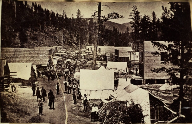 View of the main street of Deadwood, South Dakota.  Pedestrians and horse-drawn wagons are on the street lined with wood buildings and tents.
