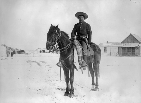 A Black Corporal, (Buffalo Soldier) in the Ninth Cavalry, poses on his horse on the Native American (Lakota Sioux) Pine Ridge Agency, South Dakota. He wears a military uniform and has a gun in a holster on his belt. Snows covers the ground and frame agency buildings show in background.