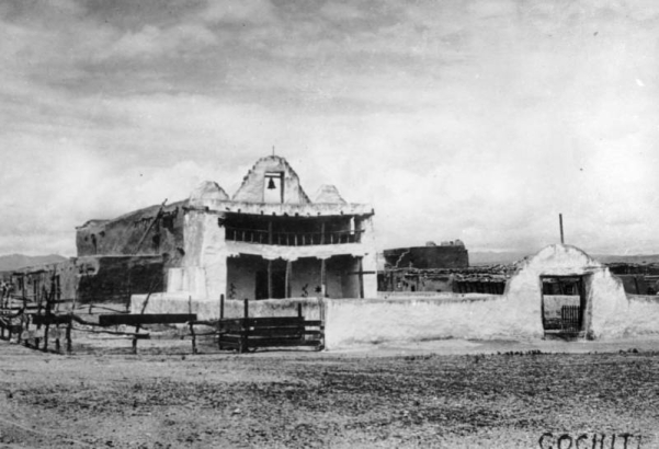 View of the Mission of San Buenaventura in Cochiti Pueblo, New Mexico, shows an adobe building with a central parapet and bell flanked by two, smaller, twin parapets, an open balcony, and an entry gate that opens to a courtyard in front of the church.