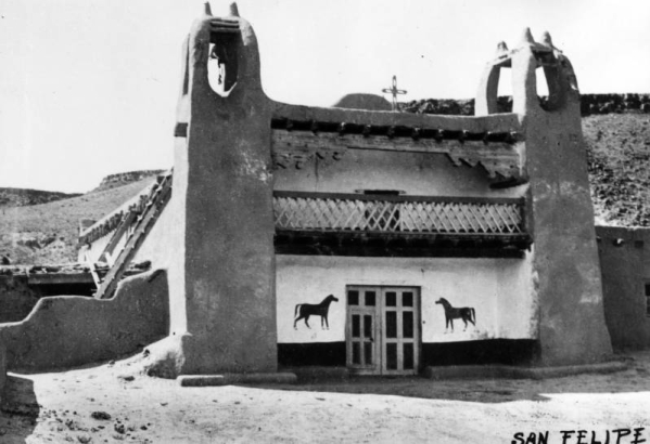 View of an adobe mission building in the Native American pueblo of San Felipe, New Mexico, shows twin bell towers, a second floor balcony, and double doors flanked by representations of horses.