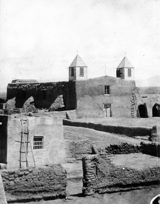 View of the Native American pueblo of Isleta, New Mexico, shows San Augustine church which features twin towers with louvered vents, adobe walls, a cross, and double doors. A ladder leans against the building; other adobe buildings nearby.
