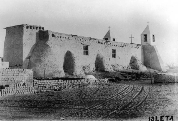 View of a mission church in the Native American pueblo of Isleta, New Mexico, shows twin bell towers with louvered vents and crosses on top, adobe wall construction, and a pile of adobe bricks.