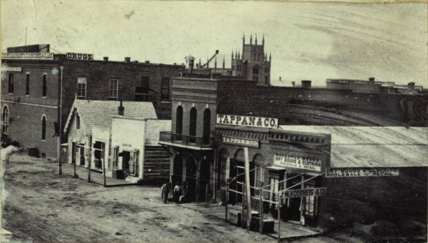 View of 15th (Fifteenth) (F) Street in Denver, Colorado. Signs on the wood and brick business district buildings read: "Broadway Store Dry Goods & Clothing Wholesale & Retail", "Tappan & Co.", "Chemicals Paints Oils Brushes & Glass", and "Drugs". Lawrence Street Methodist Episcopal Church is in the distance.