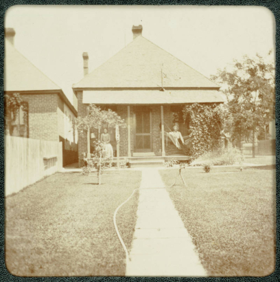 A man and woman pose on the porch of their brick home in Denver, Colorado. The lawn is watered by a hose on a stake.