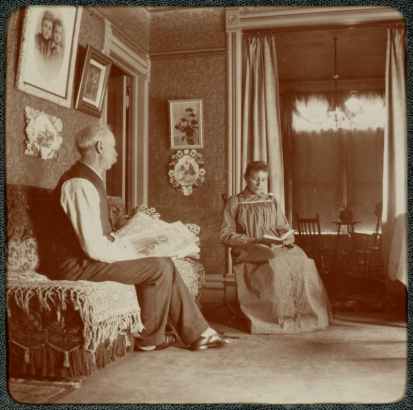 A man and woman who read in their home in Denver, Colorado. Photographs and paintings hang on the walls. The man holds a newspaper titled "Brown Book".