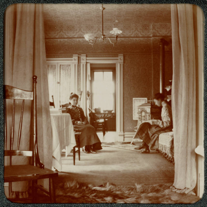 Two women sit indoors in a home in Denver. A fur carpet and a woodstove are in the house.