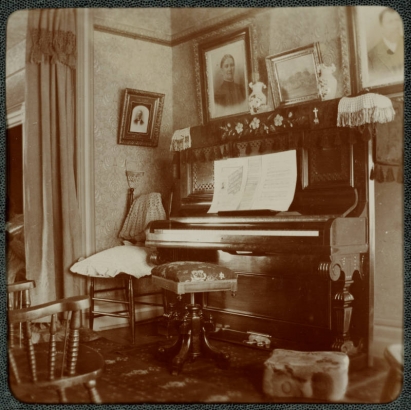 View of a piano in a house in Denver, Colorado. Sheet music titled "Where the Silvry [sic] Colorado Wends Its Way" and vases are on the piano. Photographs and paintings hang on the wall. A piano stool and chairs are nearby.