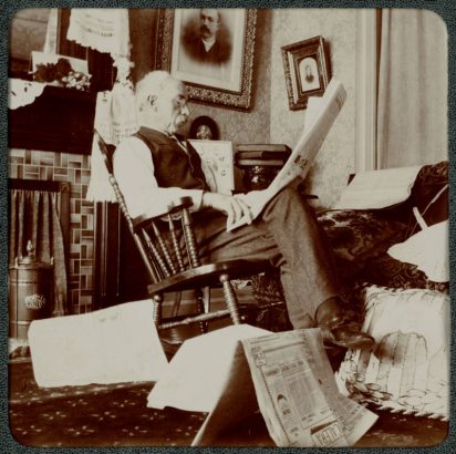 A man sits in a rocking chair and reads a newspaper in his home in Denver, Colorado. A fireplace is in the room and photographs and portaits hang on the walls. Newspapers litter the floor.