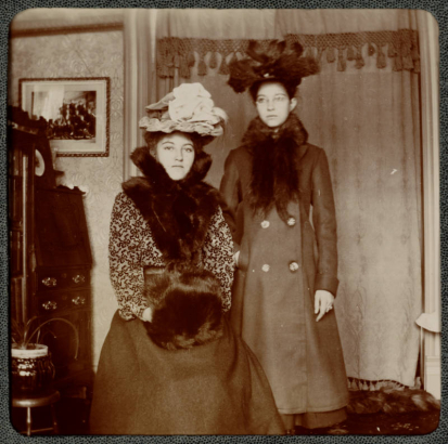 Two women pose indoors near a drop-front desk and bookcase in Denver, Colorado. One woman wears a fur muff and a hat decorated with flowers.