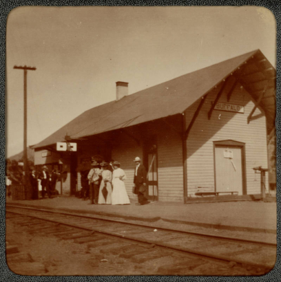 Men and women stand near the railroad depot in Buffalo, Colorado. They hold valises and umbrellas. Railroad tracks are nearby.