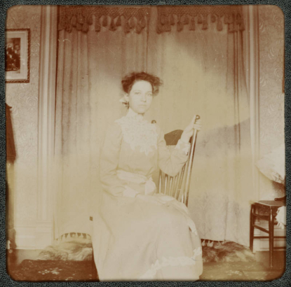 A woman poses indoors on a rocking chair in a house in Denver, Colorado. She wears a long dress.