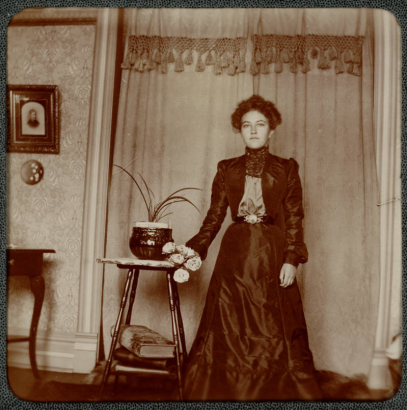 A woman poses indoors in a house in Denver, Colorado. She holds a bouquet of flowers and wears a long dress. A framed photograph hangs on the wall.